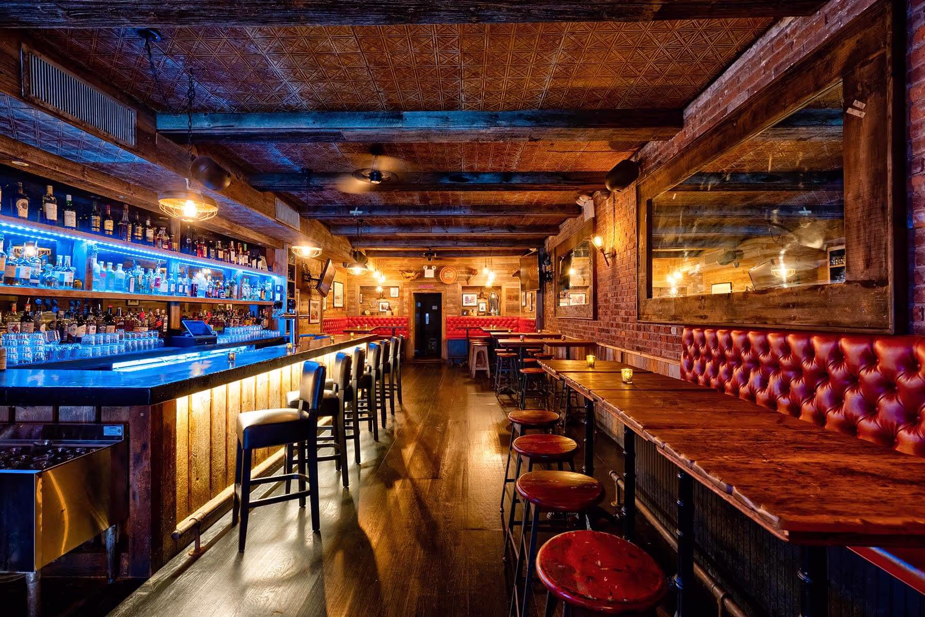 Cozy bar interior with wooden floor, long wooden bar counter with stools, red tufted booth seating, and warm ambient lighting.
